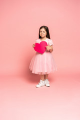 Laughing fashion little girl in tulle dress holding red paper heart and smiling to camera isolated on pink background.