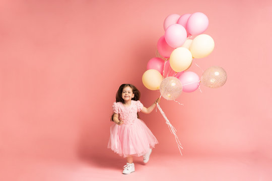 Happy celebration of birthday party with flying balloons of charming cute little girl in tulle dress smiling to camera isolated on pink background. Charming smile, expressing happiness