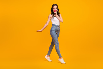 Full body profile photo of attractive cheerful lady good mood walk street arm on cheek wear white tank-top jeans footwear isolated bright yellow color background