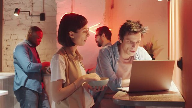 Young Caucasian woman eating snack and discussing project on laptop with male colleague while cooperating in modern loft coworking space; multiethnic men working in background