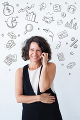 Happy woman talking on cell with hand drawn business sketches. Middle aged curly haired lady in casual standing over background with icons and speaking on mobile phone. Good news concept