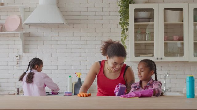Happy attractive african american mother and two cute preadolescent daughters doing cleanup using detergents in domestic kitchen. Positive little girls helping mother with household chores indoors.