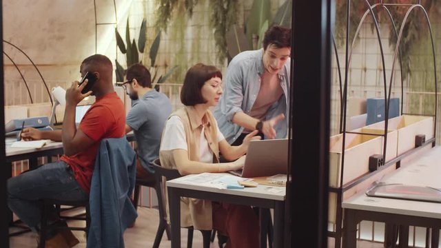 Cheerful young woman greeting male colleague in modern coworking space and discussing project on laptop with him while multiethnic men working behind them