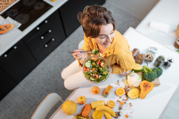 Portrait of a young cheerful woman eating salad at the table full of healthy raw vegetables and...