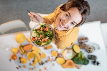 Portrait of a young cheerful woman eating salad at the table full of healthy raw vegetables and fruits on the kitchen at home, view from above. Concept of vegetarianism, healthy eating and wellness