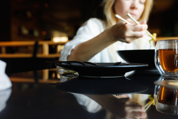Fototapeta na wymiar a girl in a Chinese restaurant eats noodles with chopsticks, on a table is a glass cup of teas and lies a smartphone