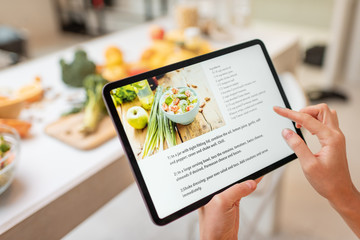 Woman looking on the digital recipe, using touchscreen tablet while cooking healthy meal on the kitchen at home, close-up view on the screen - obrazy, fototapety, plakaty