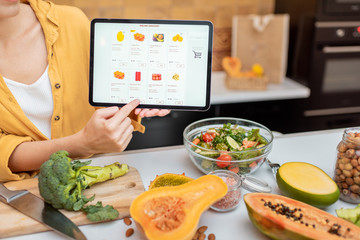 Woman holding digital tablet with launched online shopping market while standing on the kitchen with lots of fresh food on the table