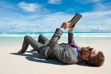 Relaxed office worker lying in a suit with his tablet on the shore of a tropical beach