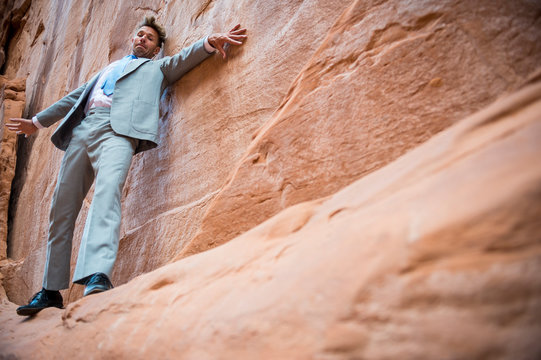 Nervous businessman clinging to a cliff face while balancing on a narrow ledge in a red rock canyon