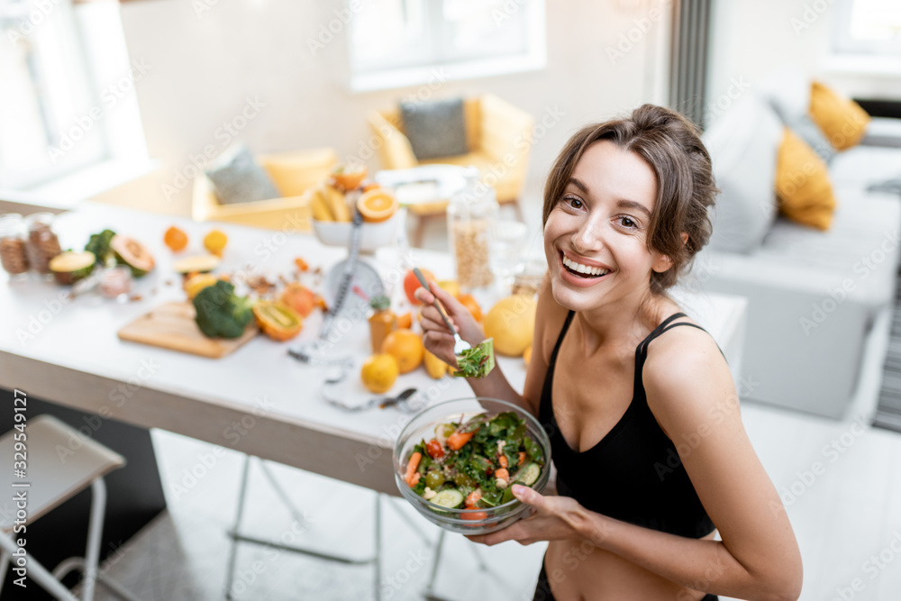 Wall mural Portrait of a cheerful athletic woman eating healthy salad during a break at home. Concept of losing weight, sports and healthy eating