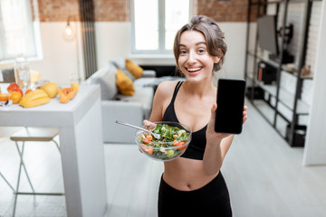 Young athletic woman showing smart phone screen, while standing with healthy food at home. Concept...