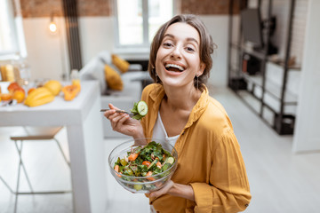 Portrait of a young and cheerful woman dressed in bright shirt eating salad at home. Concept of...