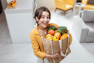 Portrait of a young and cheerful woman standing with shopping bag full of fresh fruits and...