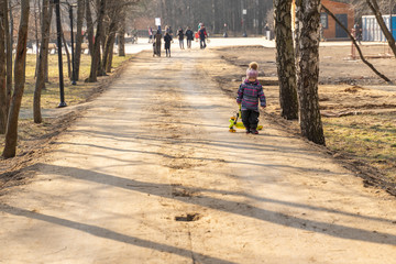 3-4 year old child in a hat and overalls walks along a dirt road in a city park and drags a scooter