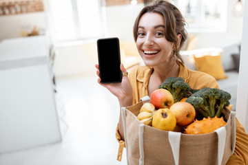 Portrait of a young and cheerful woman holding smart phone and shopping bag full of fresh fruits...