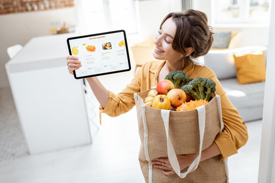 Happy woman holding a digital tablet with launched online store while standing with shopping bag full of fresh products at home. Concept of buying online using mobile devices