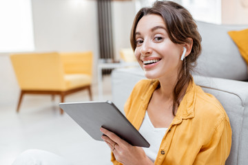 Young and cheerful woman working on digital tablet using wireless earphones while sitting relaxed at home. Concept of leisure, freelance and mobile work