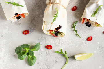 Healthy lunch snack. Three vegan tortilla wraps, rolls sandwiches with vegetable, green salad mix, lime and chili pepper on a light grey stone background. Top view.