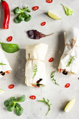 Healthy lunch snack. Three vegan tortilla wraps, rolls sandwiches with vegetable, green salad mix, lime and chili pepper on a light grey stone background. Top view.