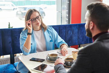 Attractive young businesswoman adjusting eyeglasses and chatting with colleague at meeting in modern cafe