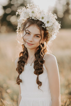 portrait of a smiling young girl in a white dress and a floral wreath on her hair with braids in the summer field