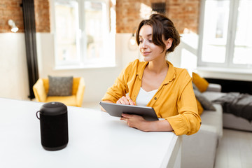 Happy young woman controlling smart home devices with a voice commands and smart speaker at home. Concept of a smart home and managing wireless devices remotely