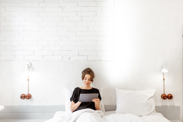 Young woman with a digital tablet lying in bed, wide interior view of a white bedroom. Concept of a leisure time with mobile wireless devices at home