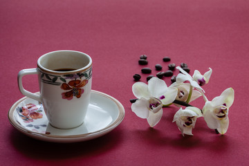 Espresso coffee in slim cup with floral pattern with orchid flowers and some spare coffee beans. Burgundy color background.