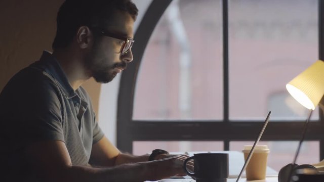 Tilt up shot of young middle eastern man in casual t-shirt and glasses typing on laptop and drinking tea from mug while working at desk near arch window