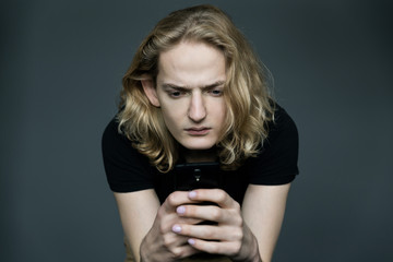 Young man with long blonde hair taking pictures of something with a phone with a strained expression on his face on a black background