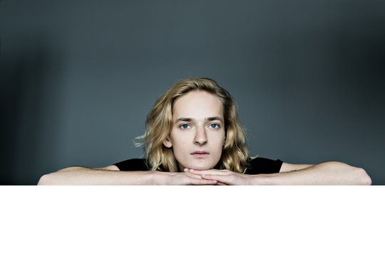 A young attractive man peeks out from above a white wall on a gray background. Looks surprised laid his head in his hands. The elbows are apart.