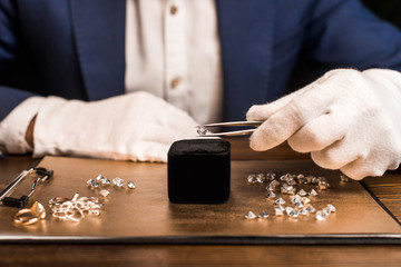 Cropped view of jewelry appraiser holding gemstone in tweezers near jewelry on board on table...