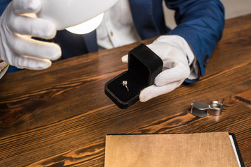 Cropped view of jewelry appraiser holding ring with gemstone in box near lamp at table isolated on...
