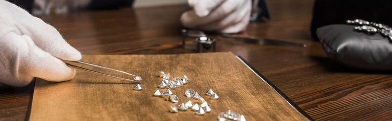 Cropped view of jewelry appraiser with tweezers examining gemstones on wooden table isolated on grey, panoramic shot