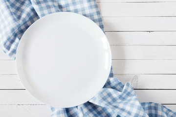 Empty white plate on wooden white table with checkered blue linen napkin. Flat lay, top view, copy...