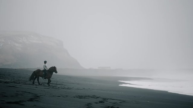 Long lens shot of a girl dressed in traditional Icelandic sweater riding horse on a black beach, Iceland