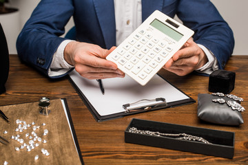 Cropped view of jewelry appraiser holding calculator near clipboard and jewelry with gemstones on...