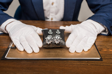 Cropped view of jewelry appraiser holding jewelry earnings on jewelry pillow near board on table...