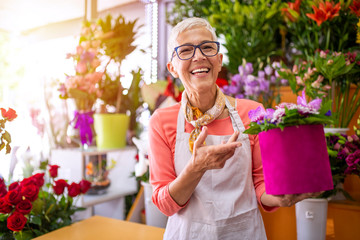 Mature smiling florist shop owner surrounded by flowers. Beautiful mature female , smiling works at flower shop. Portrait of mature female florist with flower box looking at camera - obrazy, fototapety, plakaty