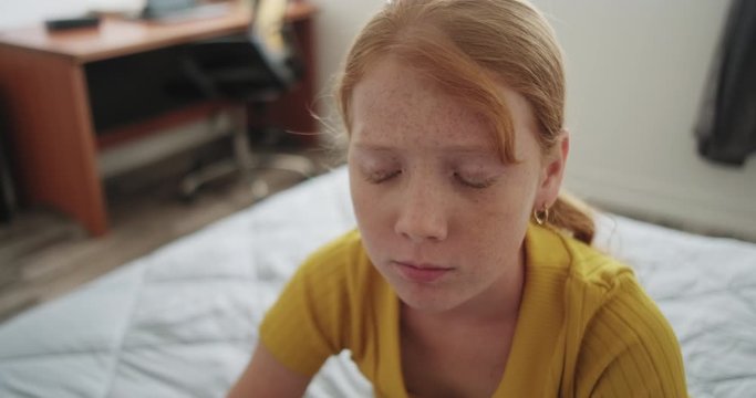 Bullied sad little girl in bedroom at home. Lone redhead female child sitting on bed with emotions, feelings of sadness and loneliness