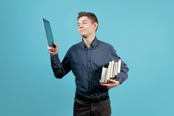 A teenager in a blue shirt holds a tablet and a stack of books under his armpit. Raises the tablet and looks joyfully at the tablet