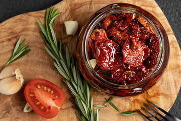 Sun-dried tomatoes with garlic, rosemary and spices in a glass jar on an olive wood cutting board, top view, flat lay
