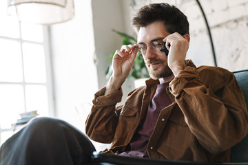 Image of unshaven young man holding eyeglasses while resting in cafe