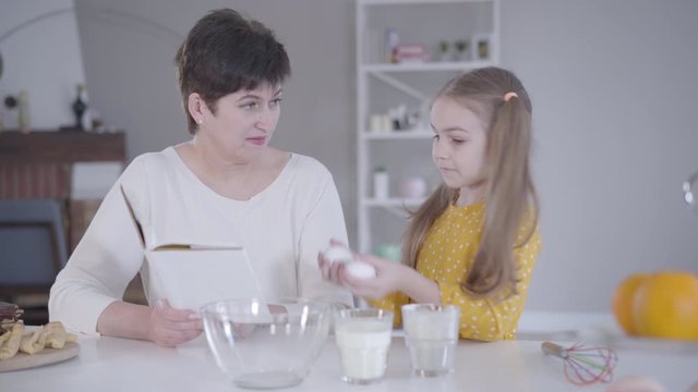 Portrait of diligent little girl helping grandmother with baking of pancakes for Shrove Tuesday. Pretty Caucasian child gathering eggs, flour and milk as woman reading ingredients from recipe.