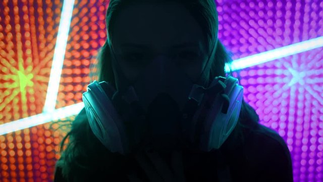 Young girl model in the neon lights of the club. Concept of dubstep or punk style. Wearing a gas mask from dirty air. Protected in a public place from the Chinese coronavirus.