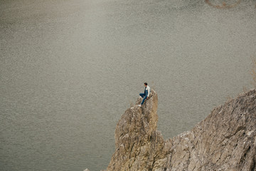 Extreme man in outdoor sitting on rock of lake. Hipster traveler sitting on top of rock and enjoying amazing view on lake. Stylish guy exploring and traveling. Atmospheric tranquil moment.