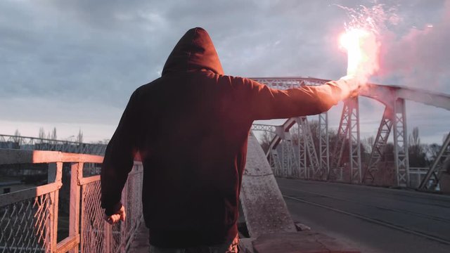 Back view of a young man in hoodie and balaclava with red burning signal flare on the road under an old steel frame bridge, slow motion