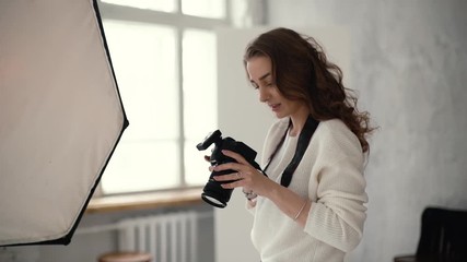 Young professional female photographer checks the pictures on the camera screen in light photo studio, on background of softbox. Concept of creative work in photo studio. Shooting in slow motion.