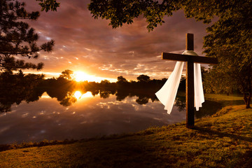 Beautiful photo illustration of an Easter morning sunrise on a cross by a calm lake. The warm light almost announces Jesus's rising from the dead.